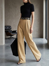Laden Sie das Bild in den Galerie-Viewer, A model wearing a black short-sleeve mock neck top and high-waisted beige wide-leg trousers with a black belt, styled with a black tote bag and black slide sandals in a modern indoor setting.
